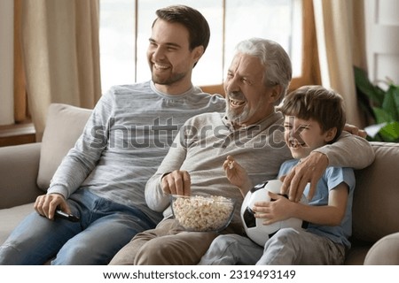 Overjoyed three generations of men relax on sofa in living room watch football match eat popcorn. Happy boy with young father and elderly grandfather have fun rest at home on family weekend together.