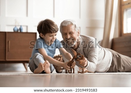 Happy elderly grandfather lying on floor at home have fun play toys with small 6s grandson together. Smiling mature grandparent feel playful engaged in funny activity with little boy child on weekend. Royalty-Free Stock Photo #2319493183