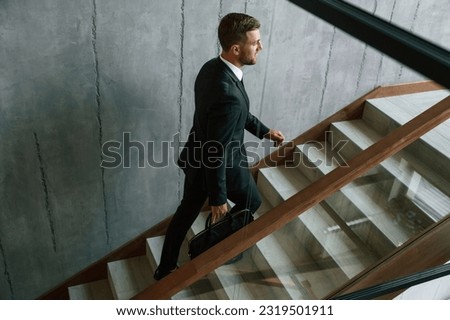 Walking up. Man in business suit and tie with case in hands is on the stairs. Royalty-Free Stock Photo #2319501911