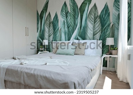 Comfortable bed with pillows in stylish bedroom in apartment. Modern wallpaper on the wall in green leaves. Design interior for relax and rest.