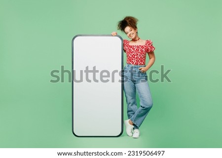 Full body smiling happy young woman she wearing casual clothes red blouse stand near big huge blank screen mobile cell phone smartphone with area isolated on plain pastel light green color background