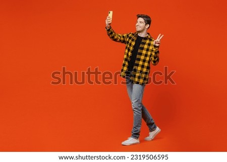 Full body young caucasian man wear yellow checkered shirt black t-shirt doing selfie shot on mobile cell phone post photo on social network show v-sign isolated on plain red orange background studio