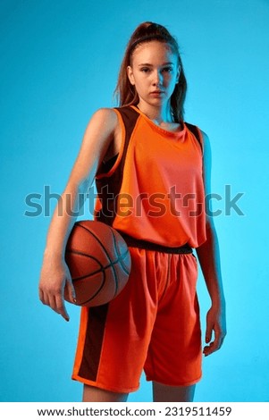 Portrait of young girl, basketball player in uniform posing with ball against blue studio background in neon light. Concept of professional sport, action and motion, game, competition, hobby, ad