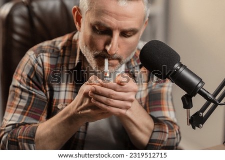 A bearded man sits at a table in front of a microphone and lights a cigarette. A gray-haired man in a plaid shirt smokes while sitting at the table.