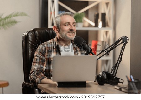 A male blogger conducts an entertaining online broadcast and communicates with his viewers. Smiling white-haired man is recording video using a studio microphone.