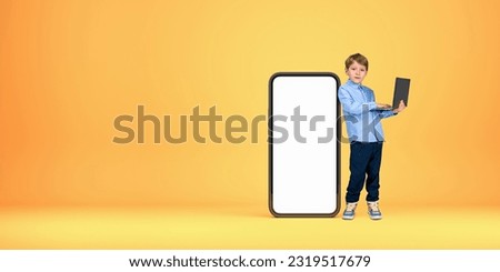 Concentrated little boy kid with laptop standing near big smartphone with mock up screen over orange background. Concept of advertising, social media and e-learning