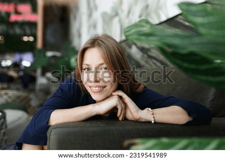 Happy young woman looking at camera. Portrait of comfortable woman relaxing on armchair. Portrait of beautiful girl smiling and relaxing on green plants background.