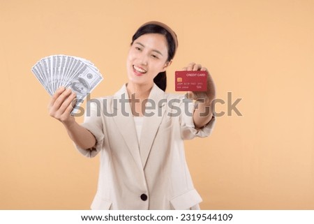portrait of successful happy confident young asian business woman wearing white jacket holding cash money dollars and credit card standing over beige background. millionaire business, shopping concept