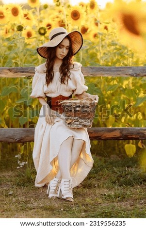 A beautiful young tall slender brunette in a beige dress and leather corset sits in a field with blooming yellow sunflowers at sunset.