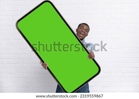 Excited black man standing and pointing Big smartphone with blank green screen over white brick background, mockup image