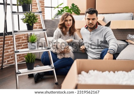 Young hispanic couple sitting on the floor at new home with log thinking attitude and sober expression looking self confident 