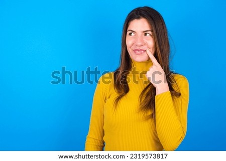 Lovely dreamy young beautiful woman wearing yellow sweater over blue studio background keeps finger near lips looks aside copy space.