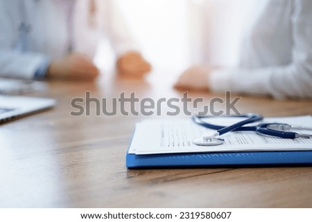 Stethoscope and medication history records form are lying on the wooden table while doctor and patient sitting and talking to each other at the background. Medicine