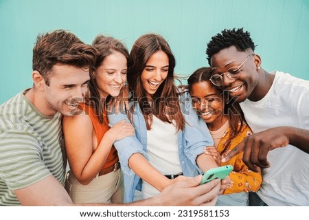Multiracial group of young friends enjoying and smiling using their mobile phone app at teal blue wall background. Diverse teenage student people having fun watching content on the social media. High