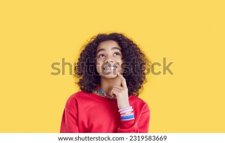 Happy little African American girl teenager looking up with smile and dreaming coming up with idea for birthday present or fun after-school vacation stands in studio on yellow background Royalty-Free Stock Photo #2319583669