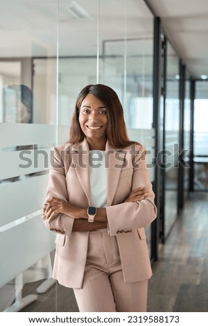 Confident smiling young professional business woman ceo corporate leader, female African American lawyer or hr manager wearing suit standing arms crossed in office, vertical portrait.
