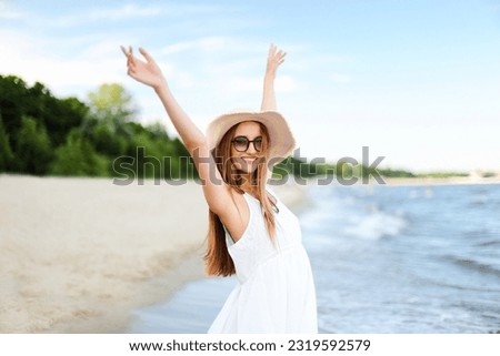 Happy smiling woman in free happiness bliss on ocean beach standing with a hat, sunglasses, and rasing hands. Portrait of a multicultural female model in white summer dress enjoying nature during Royalty-Free Stock Photo #2319592579