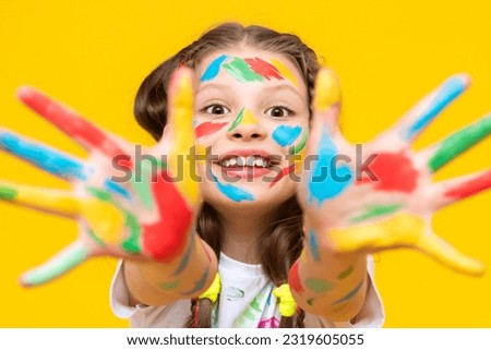 Childish pranks. A charming child with painted palms with multicolored paints. A little girl happily draws with her hands. Yellow isolated background. Royalty-Free Stock Photo #2319605055