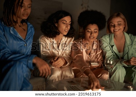 Smiling multiethnic girlfriends in colorful pajama looking at tarot cards on table near glass of wine and candles during girls night at home, bonding time in comfortable sleepwear, slumber party Royalty-Free Stock Photo #2319613175