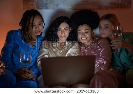 Smiling multiethnic girlfriends in colorful pajama holding glasses of wine and using blurred laptop during girls night at home, bonding time in comfortable sleepwear, slumber party Royalty-Free Stock Photo #2319613183