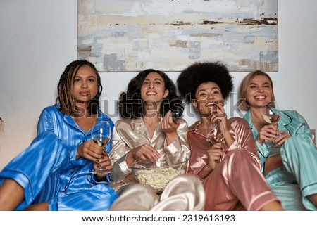Smiling multiracial women in pajama watching tv and holding popcorn and glasses of wine while sitting on bed during girls night at home, bonding time in comfortable sleepwear, slumber party Royalty-Free Stock Photo #2319613193