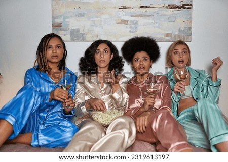 Scared multiethnic girlfriends in colorful pajama holding wine and popcorn while watching tv and sitting on bed during girls party at home, bonding time in comfortable sleepwear, slumber party Royalty-Free Stock Photo #2319613197