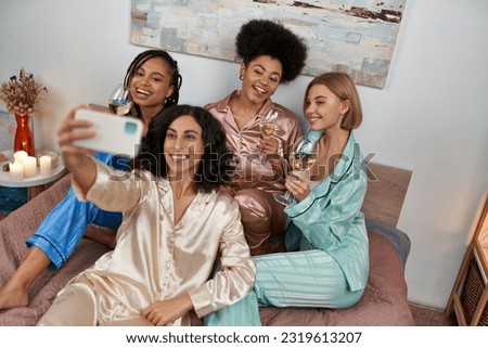 High angle view of multiracial woman in pajama taking selfie with cheerful multiethnic girlfriends with wine and sitting on bed at home, bonding time in comfortable sleepwear, slumber party Royalty-Free Stock Photo #2319613207