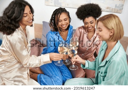 Positive and multiethnic girlfriends in colorful pajama toasting with glasses of wine while sitting on bed during girls night at home, bonding time in comfortable sleepwear, slumber party Royalty-Free Stock Photo #2319613247