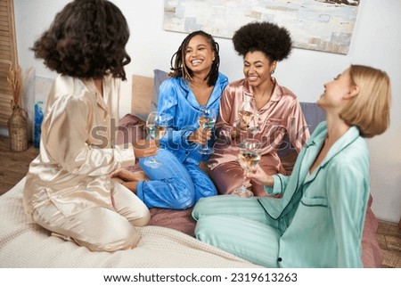 Cheerful and multiethnic girlfriends in colorful pajama holding glasses of wine and talking while sitting on bed during girls night at home, bonding time in comfortable sleepwear, slumber party Royalty-Free Stock Photo #2319613263