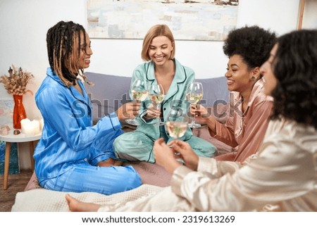 positive and multiethnic girlfriends in colorful pajama holding glasses of wine while talking during girls night on bed at home, bonding time in comfortable sleepwear, slumber party Royalty-Free Stock Photo #2319613269