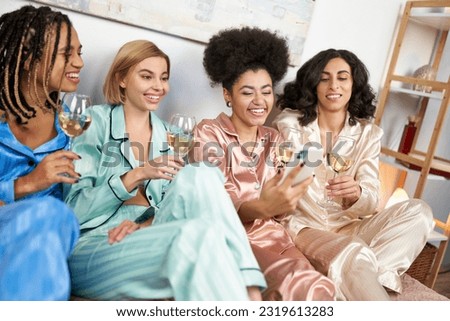 Positive and multiethnic girlfriends in colorful pajama using smartphone and holding glasses of wine while sitting on bed during girls night at home, comfortable sleepwear, slumber party Royalty-Free Stock Photo #2319613283