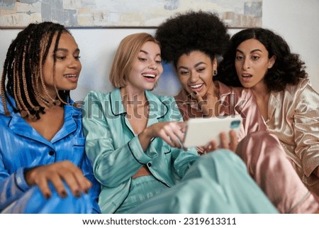 Excited blonde woman using blurred smartphone and sitting near cheerful multiethnic girlfriends in colorful pajama during girls night at home, bonding time in comfortable sleepwear, slumber party Royalty-Free Stock Photo #2319613311