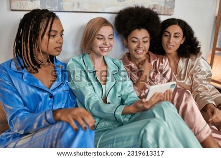 Smiling blonde woman using and holding smartphone near multiethnic girlfriends in colorful pajama during pajama party at home, bonding time in comfortable sleepwear, slumber party Royalty-Free Stock Photo #2319613317