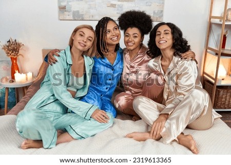 Cheerful and multiethnic girlfriends in colorful pajama hugging and looking at camera while sitting on bed during pajama party at home, bonding time in comfortable sleepwear, slumber party Royalty-Free Stock Photo #2319613365