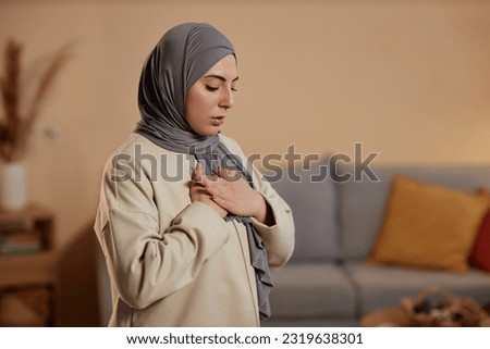 Young Islamic woman in grey hijab and beige sweatshirt expressing gratitude to her God during pray while keeping her hands on chest