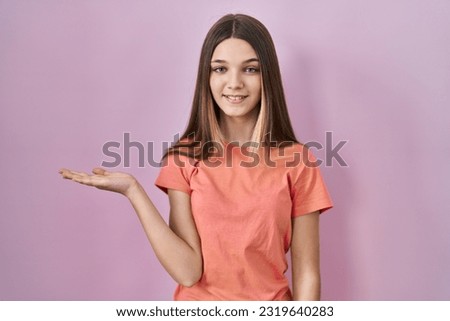 Teenager girl standing over pink background smiling cheerful presenting and pointing with palm of hand looking at the camera. 