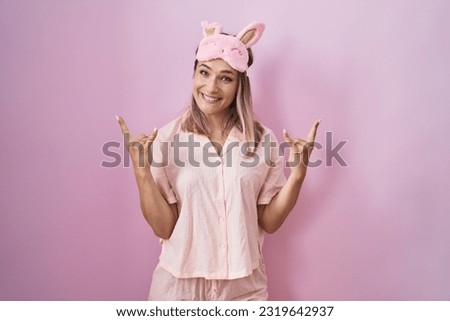 Blonde caucasian woman wearing sleep mask and pajama shouting with crazy expression doing rock symbol with hands up. music star. heavy music concept. 
