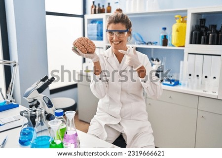 Young woman working at scientist laboratory holding brain smiling happy pointing with hand and finger 