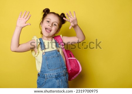 Cheerful preschooler girl showing at camera her hands palms up, smiling, enjoying the start of new school year, posing with pink backpack, dressed in blue denim dress, isolated over yellow background Royalty-Free Stock Photo #2319700975