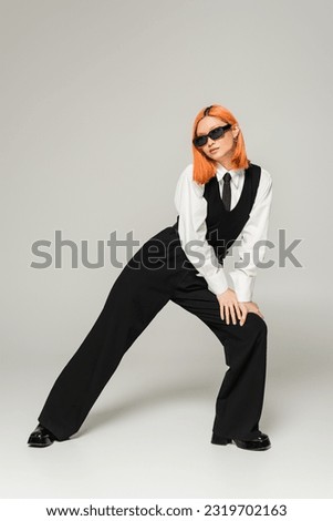 expressive and young asian woman in dark sunglasses standing in stylish pose on grey background, colored red hair, business casual, black and white clothes, generation z lifestyle, full length