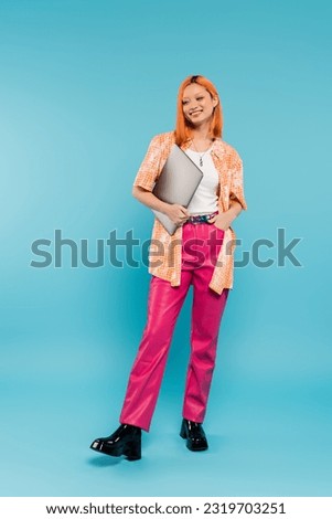 happiness and optimism, full length of red haired asian woman in orange shirt holding laptop and standing with hand in pocket of pink pants on blue background, young freelancer, generation z Royalty-Free Stock Photo #2319703251