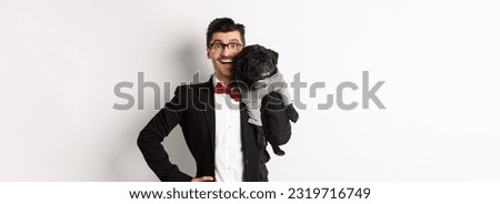 Handsome young man in suit and glasses holding cute black pug dog on shoulder, smiling happy at camera, wearing party outfits, white background.