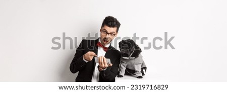 Amazed man showing video to dog, pointing at mobile phone and look excited while pug sitting unbothered in costume, white background.