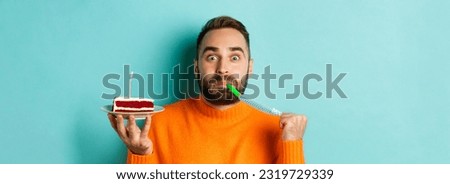 Close-up of funny adult man celebrating his birthday, holding bday cake with candle, blowing party wistle and rejoicing, standing over light blue background.
