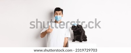 Covid-19, animals and quarantine concept. Handsome young man and small dog wearing face masks, owner showing thumb up in approval, praise super cool product, white background.