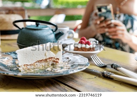 Cheesecake, strawberry tarts and tea on a table in a summer cafe. In the background is a girl who takes pictures of food on a smartphone.