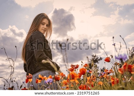 Atractive long haired  brunette young woman is looking over her shoulder at the camera in a field of red poppies. The photo is edited as a cinema photo.