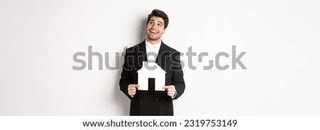 Portrait of handsome man in suit searching for home, holding paper house and looking at upper right corner dreamy, standing over white background.