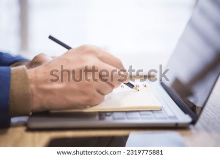 In close view, a man's hand writing down notes in a notepad on a laptop, against a hazy backdrop