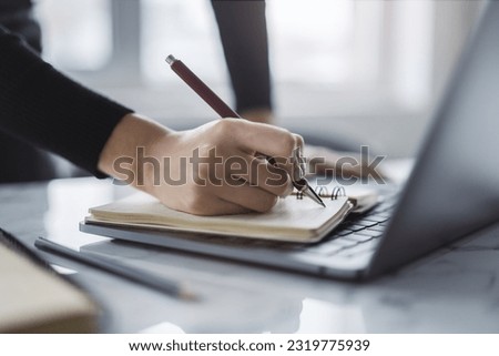 Close-up shot of a woman's hand writing in a notepad on a modern laptop, with a blurred background